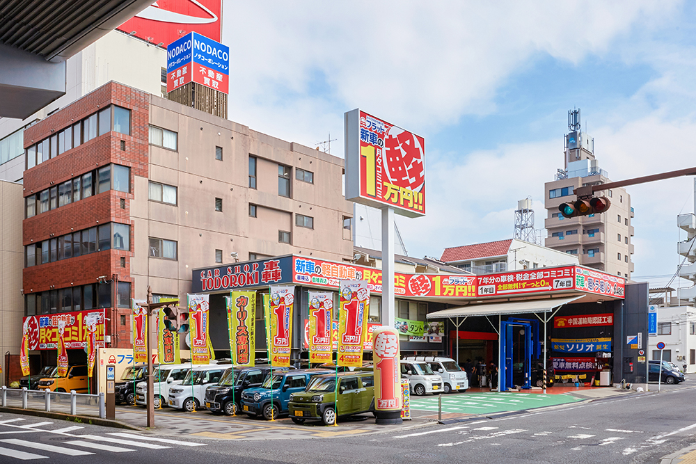 轟自動車フラット7観音の店舗外観。「新車の軽自動車が月々コミコミ1万円」の看板と、店頭に並ぶ展示車両