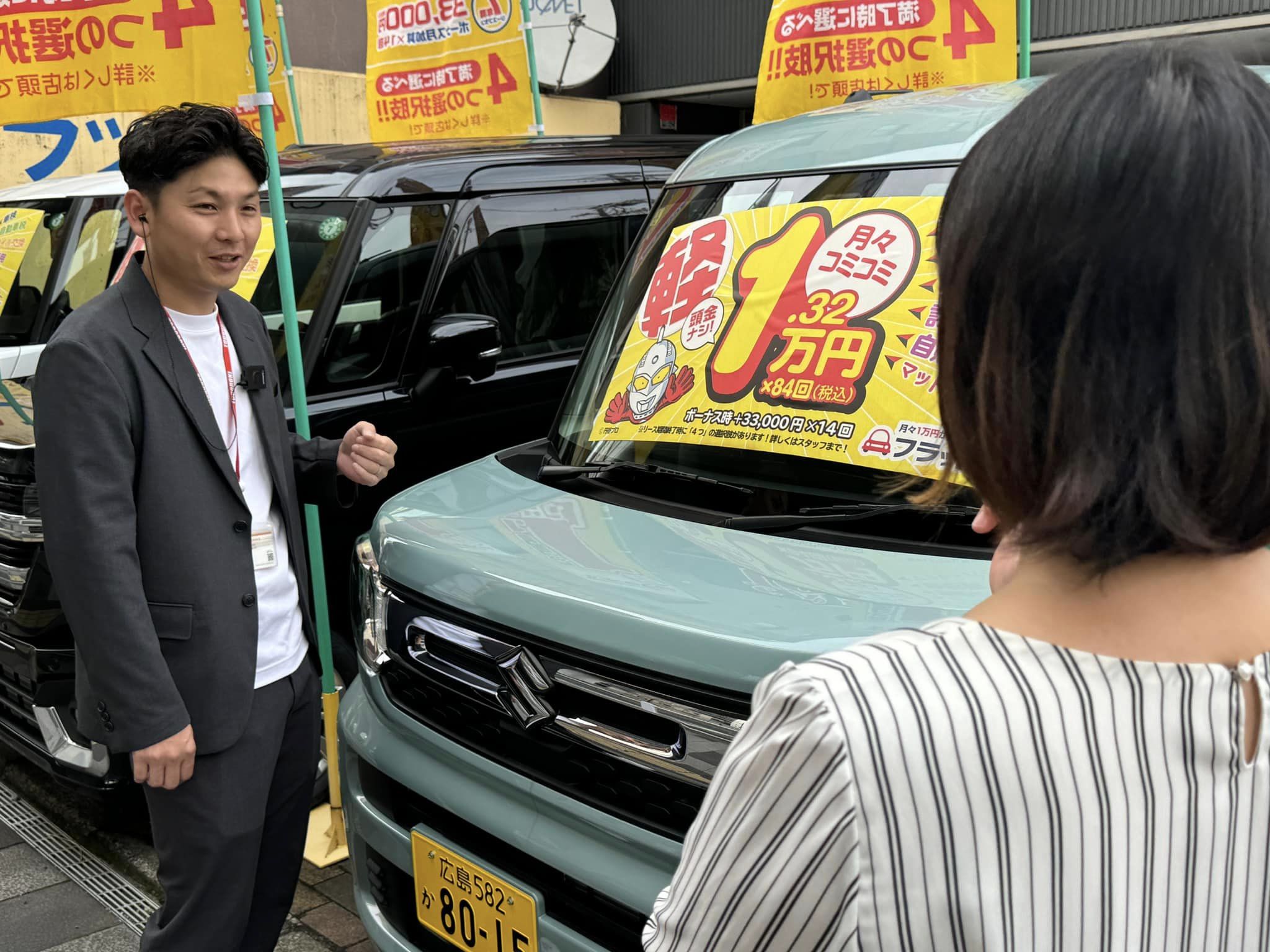 轟自動車フラット7観音のスタッフが、展示車両の前でお客様に「月々コミコミ1.32万円」プランを案内している様子
